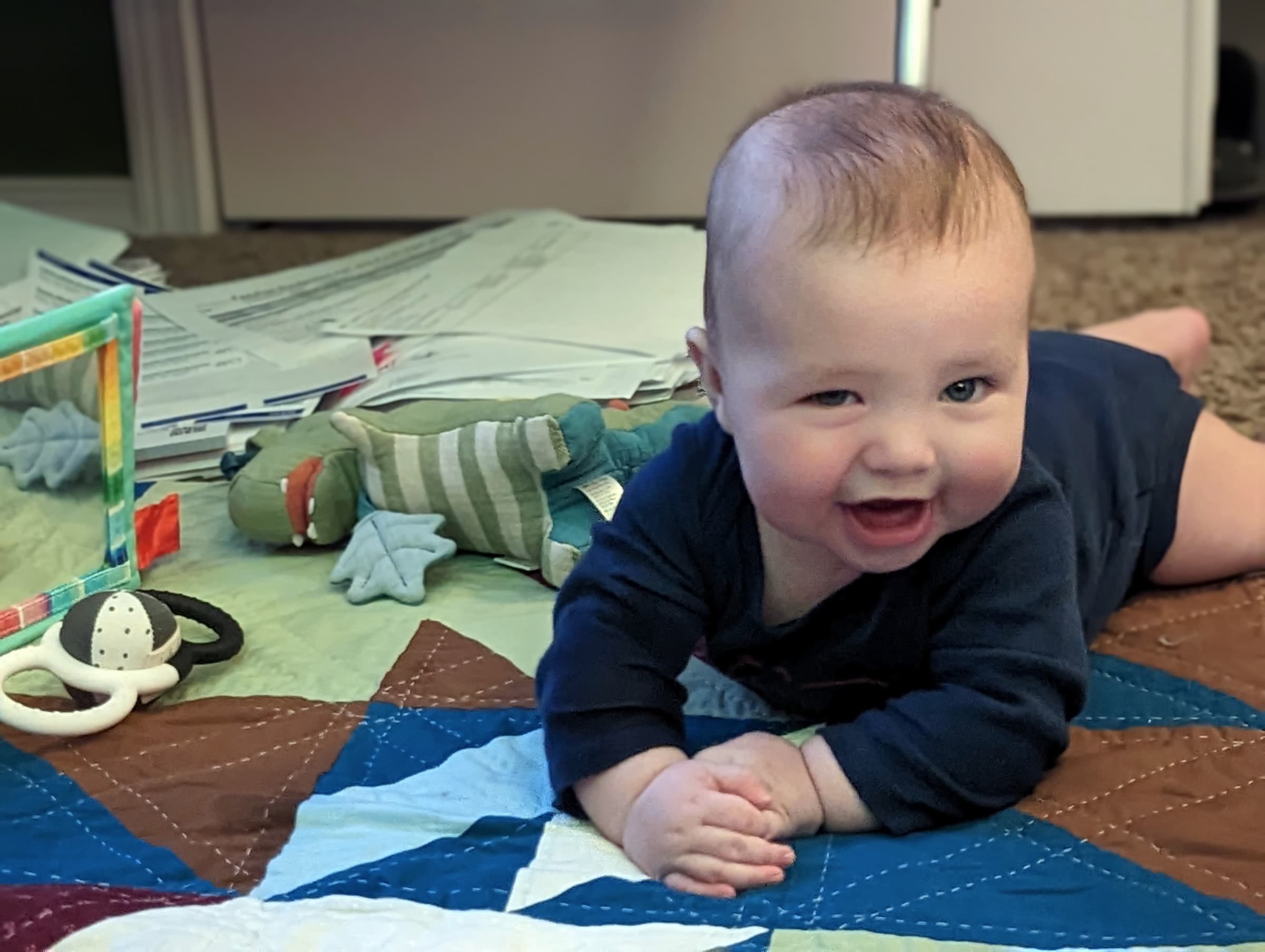 A baby laying on a quilt, looking at the camera with a goofy grin.